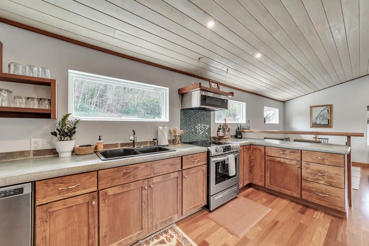 A bright, open interior showcases a modern kitchen with a central island, stainless steel appliances, and warm wooden cabinetry. Large windows flood the space with natural light, revealing a glimpse of the dining and living area beyond. The overall design blends rustic textures with contemporary comfort, offering an inviting space for cooking, gathering, and entertaining.