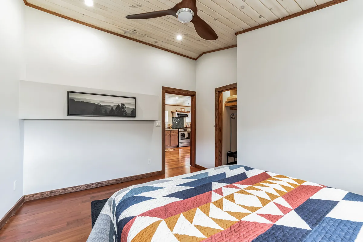 A welcoming bedroom with a colorful quilt topped bed, modern ceiling fan, and framed wall art. The room radiates comfort, with gentle lighting and a layout designed for relaxation and quiet moments—perfect for unwinding or reading.