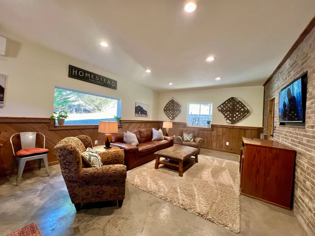 The image shows a warm, inviting living space with a plush leather sofa facing a stylish coffee table. Decorative wall art personalizes the area, while a large window fills the room with natural light and offers a glimpse of the surrounding mountain landscape. The overall ambiance is cozy, perfect for intimate family gatherings or relaxing after a day of exploring nearby wineries, hiking trails, and Hendersonville attractions.