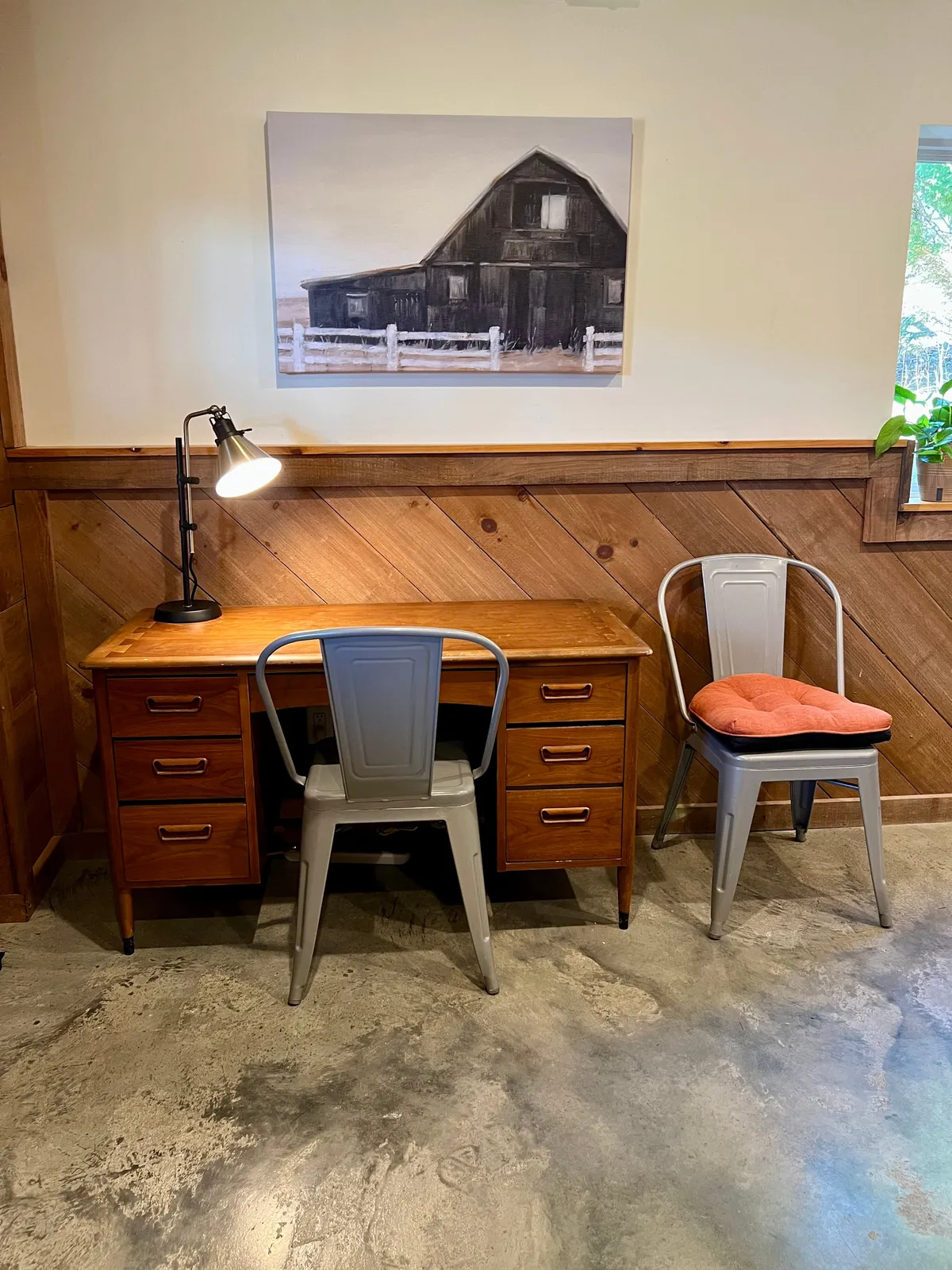 A bright and airy main-level space featuring a wooden desk with metal chairs, complemented by a tasteful artwork of a barn. The room conveys a productive yet serene atmosphere, ideal for remote work or creative pursuits. Natural light streams in, enhancing the sense of focus and calm.
