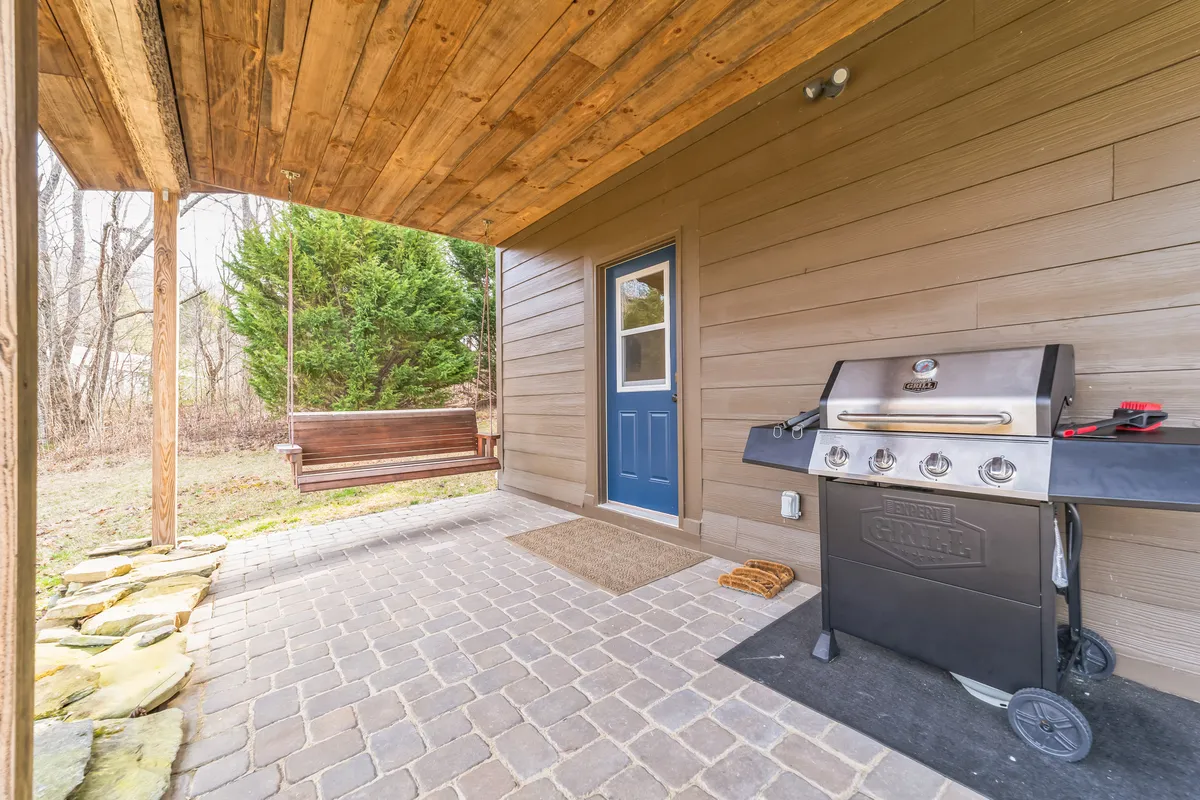This image features the front porch with a prominent propane grill, a cozy swing bench, and a welcoming blue door visible in the background. The porch setting opens to views of fresh greenery, suggesting a seamless connection to nature and outdoor spaces.