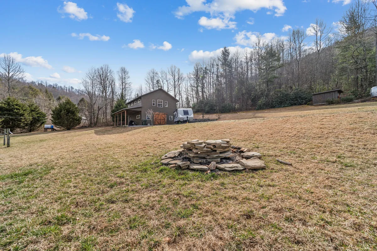 An exterior shot capturing a spacious lawn with a stone fire pit, framed by the house and surrounded by mature trees. The tranquil setting suggests opportunities for outdoor gatherings, relaxation, and exploration beyond the immediate porch area.