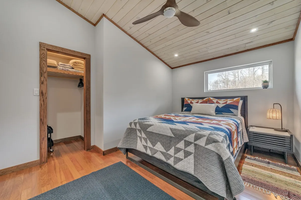 This image features a cozy bedroom with a Queen bed dressed in a patterned blanket. A nightstand with a softly glowing lamp sits beside the bed, while a window with a leafy plant provides natural light and a view outside. The closet door is visible, hinting at generous storage space.
