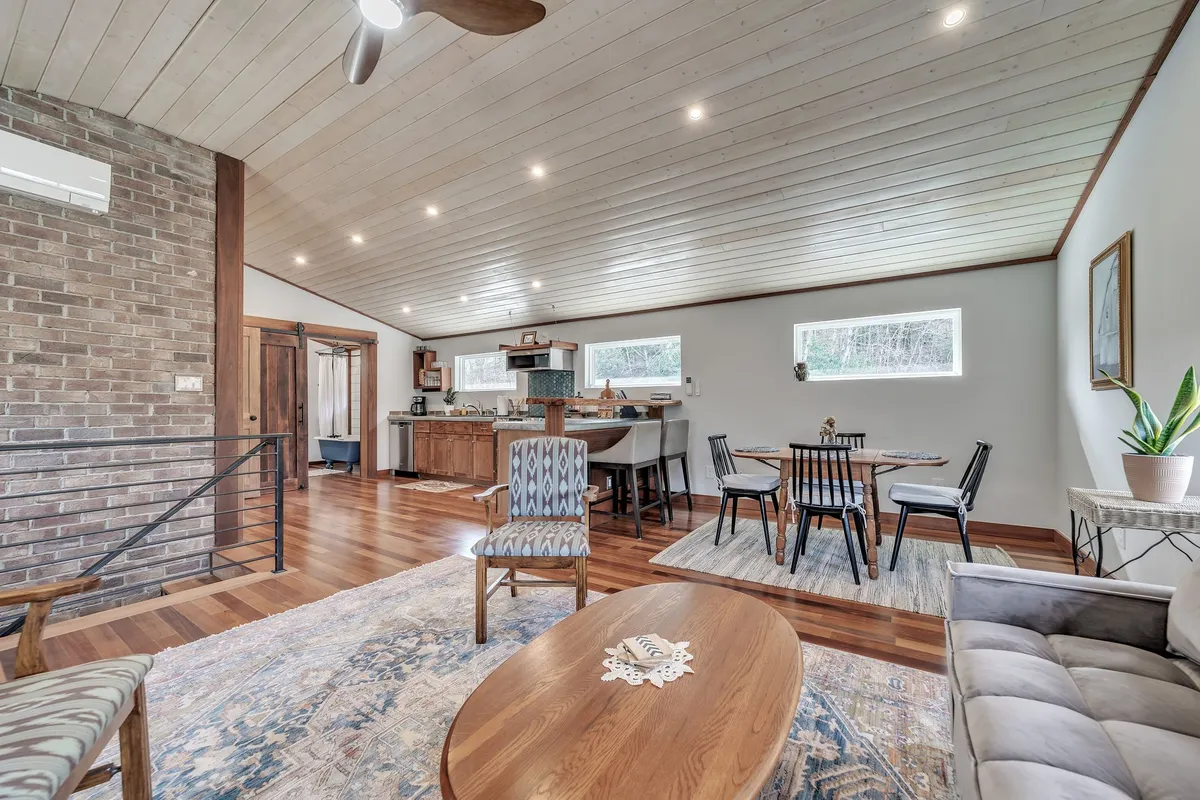 A wide angle of the living, dining, and kitchen space reveals a seamless flow between these areas. The wooden coffee table, cozy armchairs, spacious dining table, and modern kitchen are all visible, with soft daylight spilling across the room. Thoughtful decor and comfortable furniture invite relaxation and socialization.