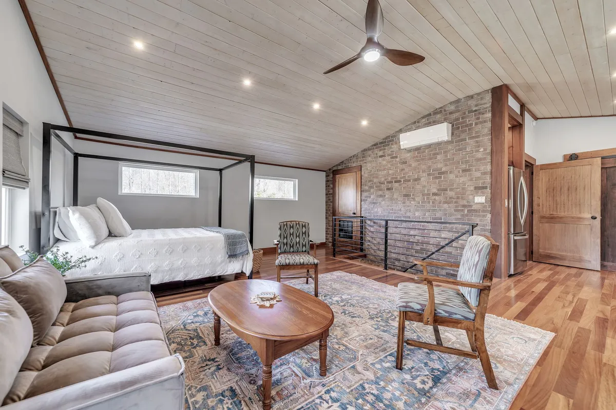 A spacious, open-plan living area featuring a cozy canopy bed, a sofa set against a rustic brick wall, and a central coffee table. Natural light fills the space, and the decor balances rustic textures with modern comfort, inviting relaxation and togetherness.